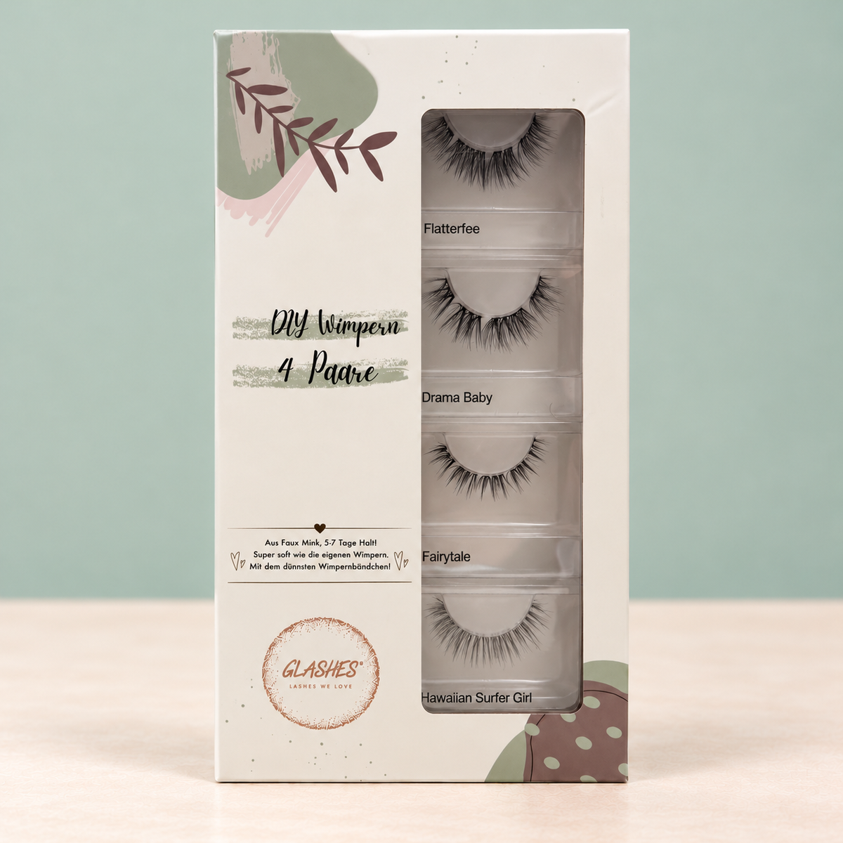 Stehende Glashes Soft Glam Box mit vier Dolleye-Wimpernpaaren vor salbeigrünem Hintergrund